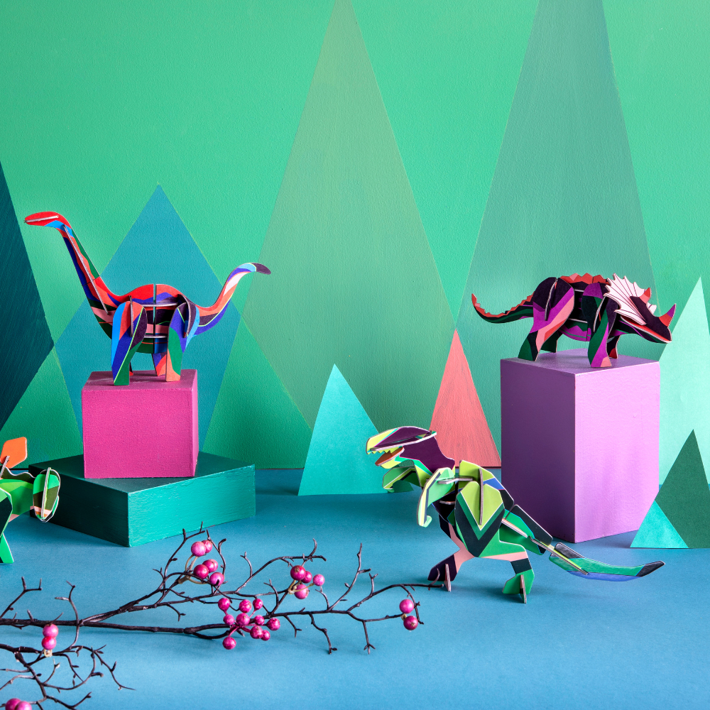 mehrere Dino 3D Deko Objekte verschiedene bunte Dinos, hintergund in sattem Grün und pink, lila Tönen, geometrische Formen