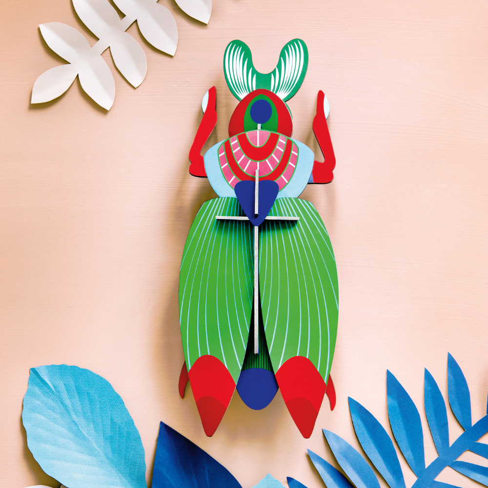 Goliathkäfer Wanddekoration dreidemensional, Wanddeko aus Karton (Recyclingkarton), bedruckt mit intensiven leuchtenden Farben, Hauptfarben: Grün, Rot, Blau. Golaithkäfer angerichtet vor pfirsich farbener Wand mit weiteren Dekoalelementen, exotische Pflan