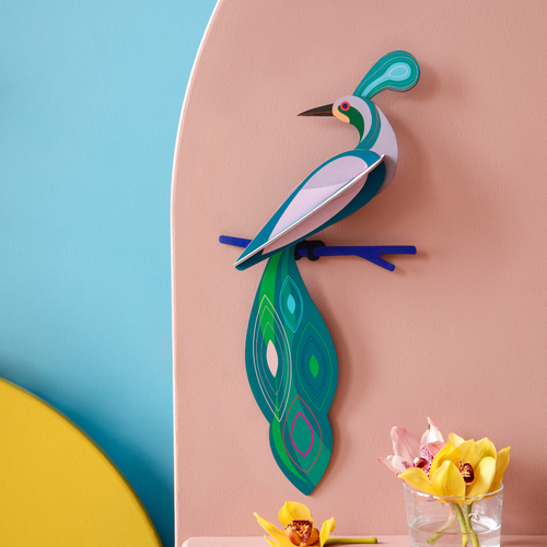 Paradiesvogel Fiji in blau, grünen edlen Tönen angeirchtet vor pfirsich farbener Wand, ein paar Blumen im Vordergrund