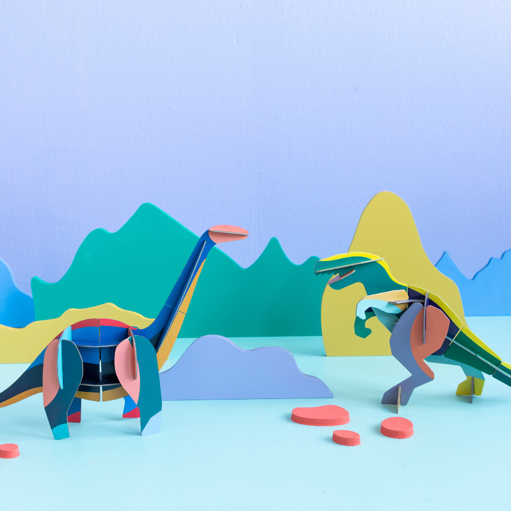 Großer Diplodocus 3D Deko Objekt zusammen mit Trex Deko Objekt in Grün gelblichen Farben angerichtet vor blau grünem Hintergrund mit farbigen Bergen aus Karton 