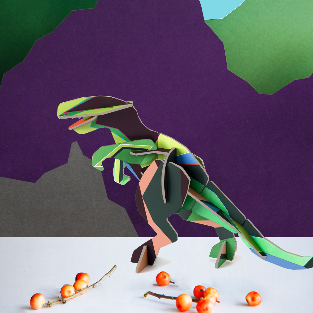 Grüner T-Rex 3D Deko Objekt aus REcyclingkarton zum zusammenstecken, farbenfroh und intensiv, angerichtet vor Hintergrund mit Bergen aus Karton, dunkellila und grün