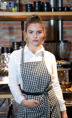 Frau trägt Schwarz/Weiß karierte Kord-Schürze in einem Coffeeshop und hält die Hand in der Schürzentasche