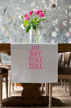 Geschirrtuch mit der Pinken Schrift "Du bist voll toll" hängt über einer TIschkante mit Blumen drauf