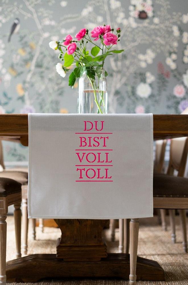 Geschirrtuch mit der Pinken Schrift "Du bist voll toll" hängt über einer TIschkante mit Blumen drauf