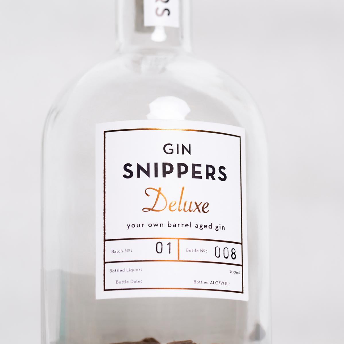 Eine mit Holzschnipsel von alten schottischen Fässern gefüllte, sonst leere GIN-Flasche mit Kork-Verschluss mit der Aufschrift "GIN SNIPPERS Deluxe" auf einem Holzbrett und daneben liegenden Schnipsel